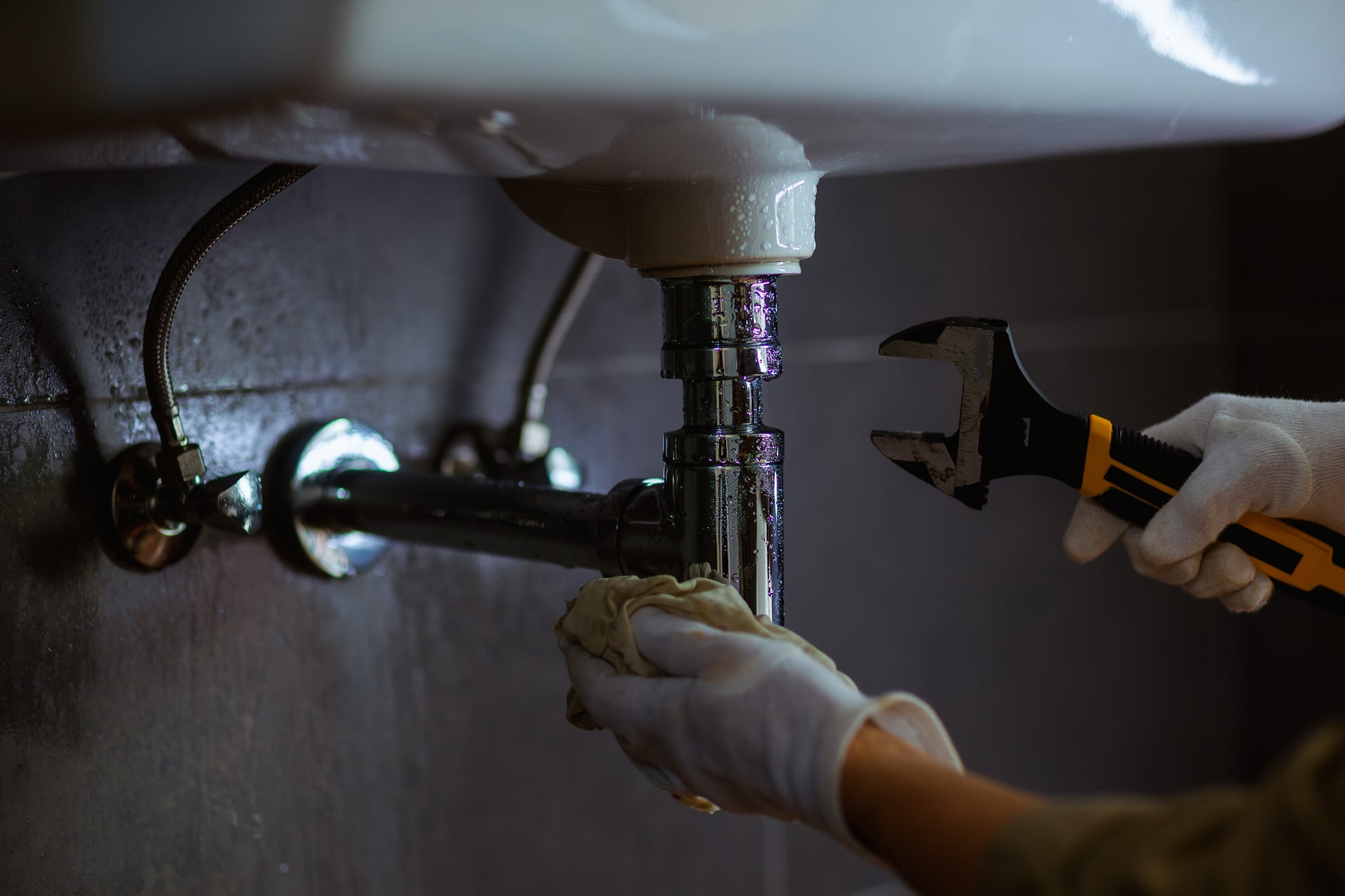 Plumber wearing protective gloves uses an adjustable wrench to repair a leaking drainpipe beneath a bathroom sink.