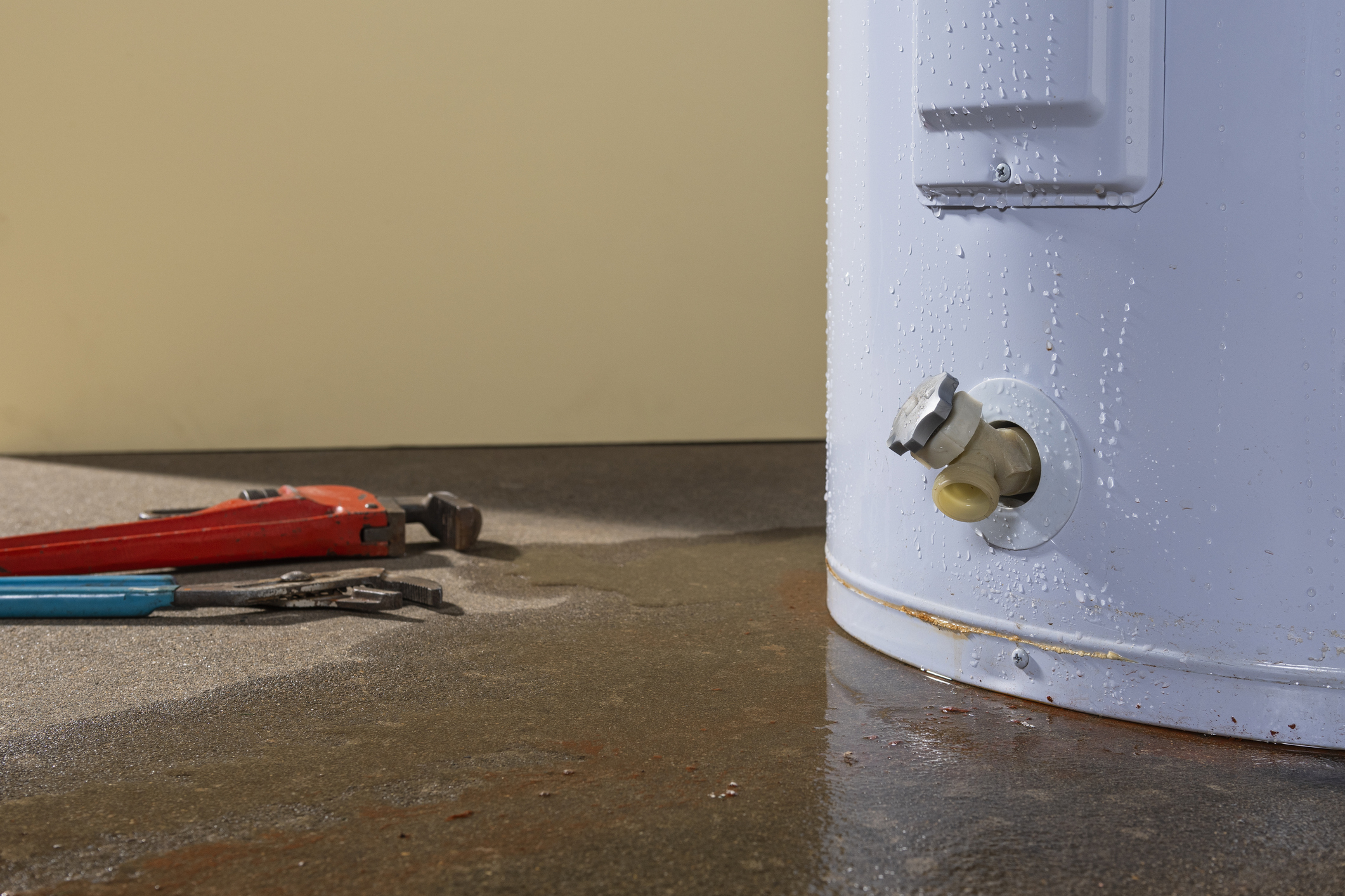Leaking water heater with water pooling on the floor and plumbing tools nearby, indicating a need for water heater repair or replacement.