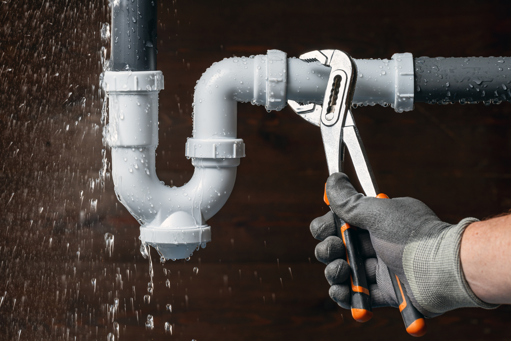 Plumber wearing work gloves uses adjustable pliers to repair a leaking PVC pipe as water sprays from the connection.